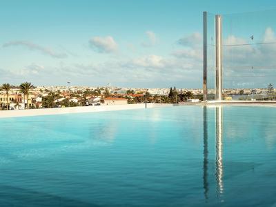 Schwimmbad mit glattem Wasser, Palmen und Stadt im Hintergrund bei blauem Himmel.
