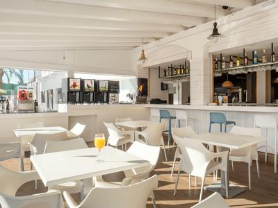 Área luminosa del bar del hotel con mesas y sillas blancas bajo techo de madera blanco.