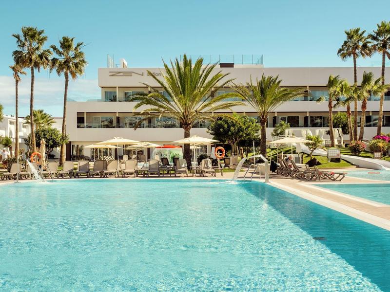 Gran piscina al aire libre con palmeras frente a un hotel blanco de varios pisos bajo un cielo despejado.