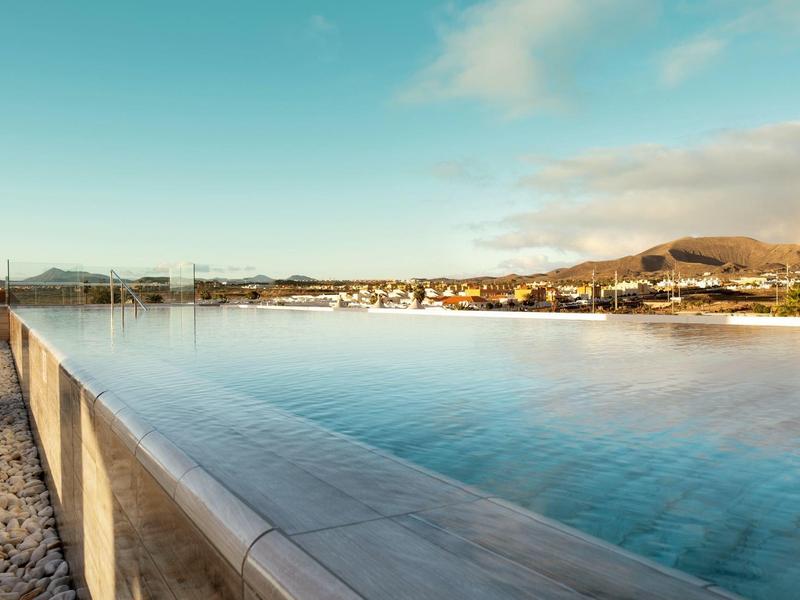 Una piscina infinita con vista a una ciudad y colinas bajo un cielo azul.