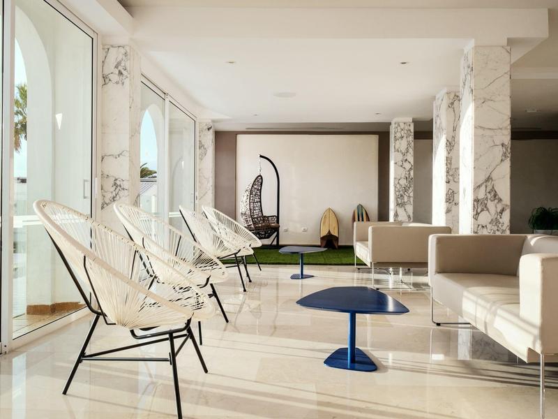 Área de salón luminosa con sillones blancos, sofás y mesas auxiliares azules frente a grandes ventanas.