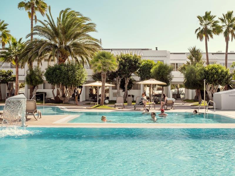 Piscina del hotel con agua clara, rodeada de palmeras y tumbonas bajo sombrillas durante el día.