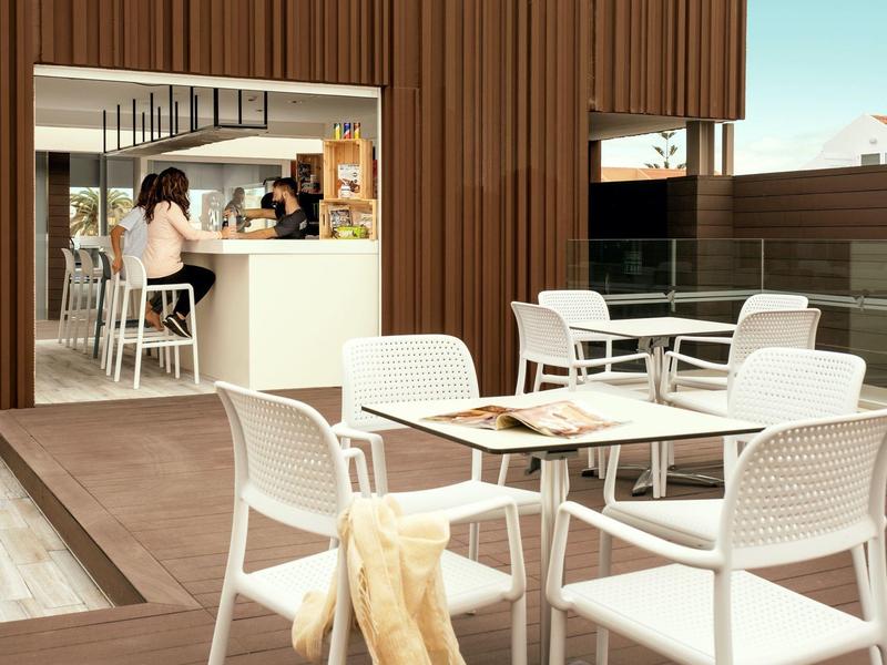 Área exterior moderna de una cafetería con mesas y sillas blancas junto a una barra.