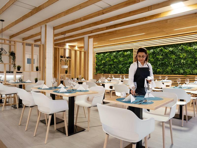 Restaurante moderno y luminoso de hotel con techo de madera, sillas blancas y decoración de pared verde.