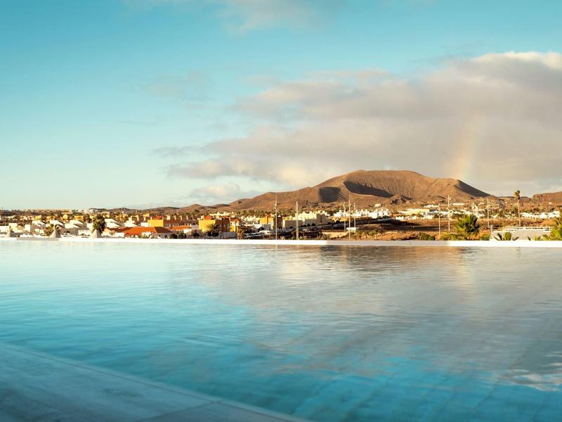 Vista de una piscina tranquila con una ciudad costera y montañas al fondo bajo un cielo despejado.
