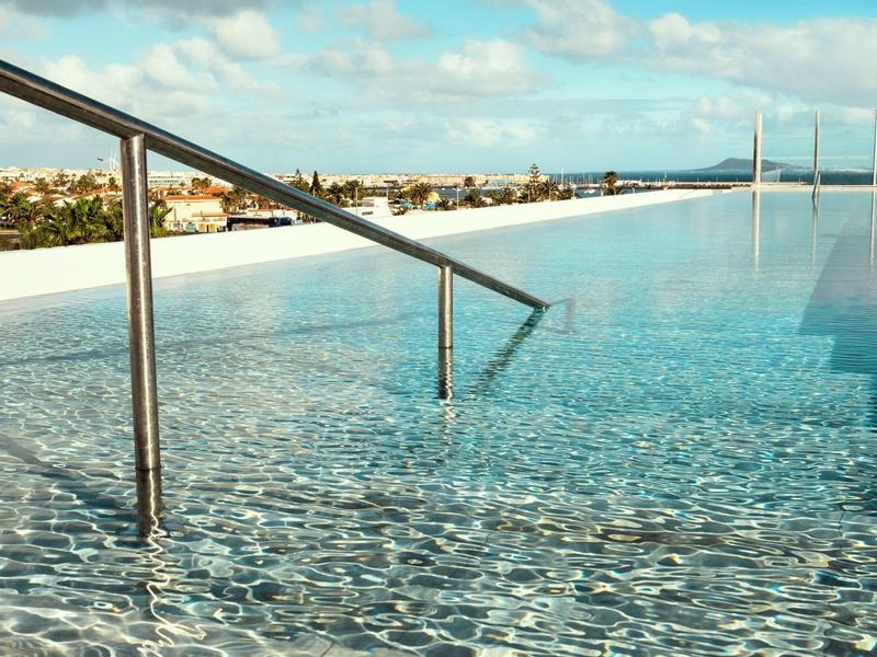Piscina infinita con agua clara y pasamanos, vista de la ciudad y el cielo al fondo.