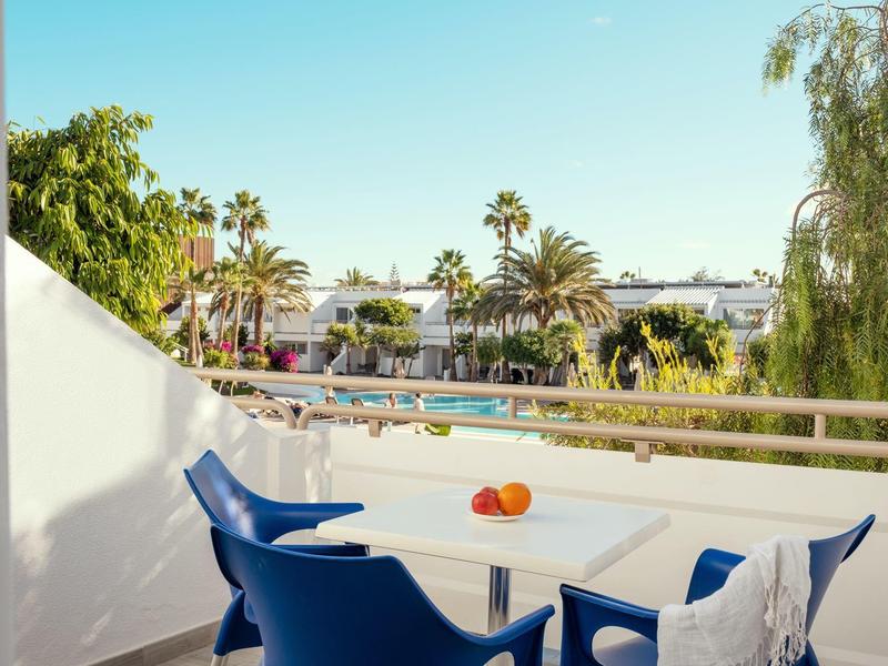 Balcón con mesa y sillas azules con vista a palmeras y casas bajo un cielo despejado.