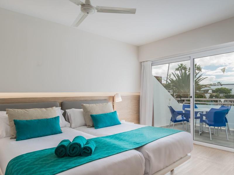 Habitación de hotel luminosa con dos camas y acceso a balcón con vista a palmeras y mar.