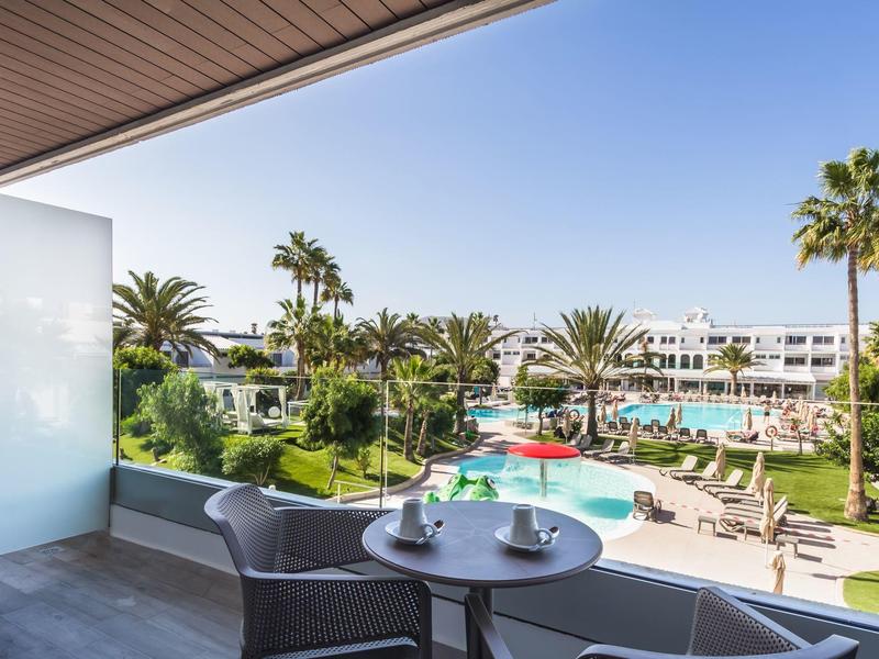 Balcón con mesa y sillas con vista a una piscina y palmeras en un resort hotelero.