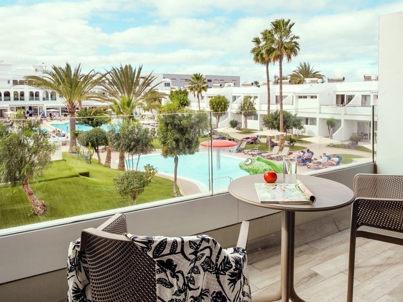 Balcón con mesa y sillas con vista a una piscina y palmeras en un complejo hotelero.
