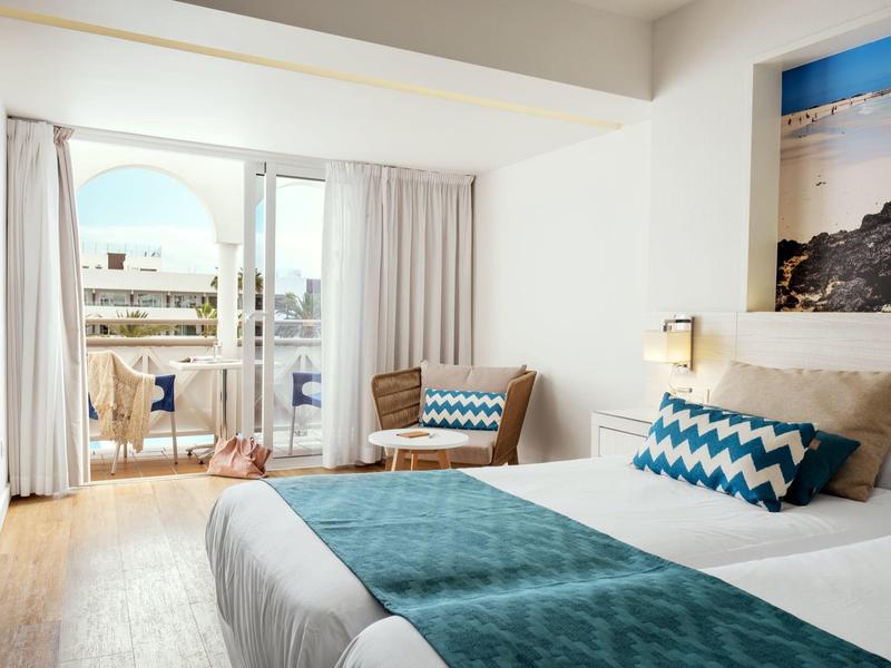 Habitación de hotel luminosa con cama doble, toques turquesa y balcón con vista.