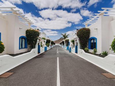 View of an empty street flanked by white buildings with blue windows and green shrubs under a blue sky.