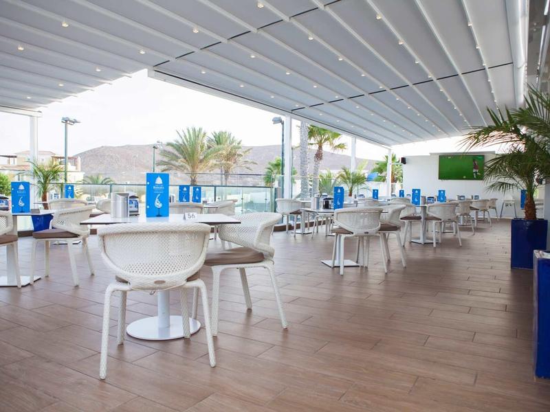 Modern open dining area with white tables and chairs, covered and overlooking palm trees.