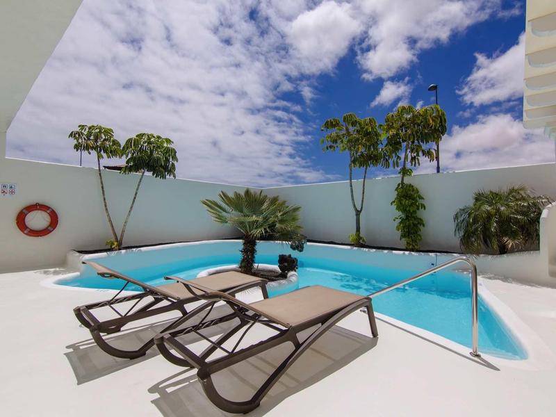 Modern pool area with sun loungers and tropical plants under a blue sky.