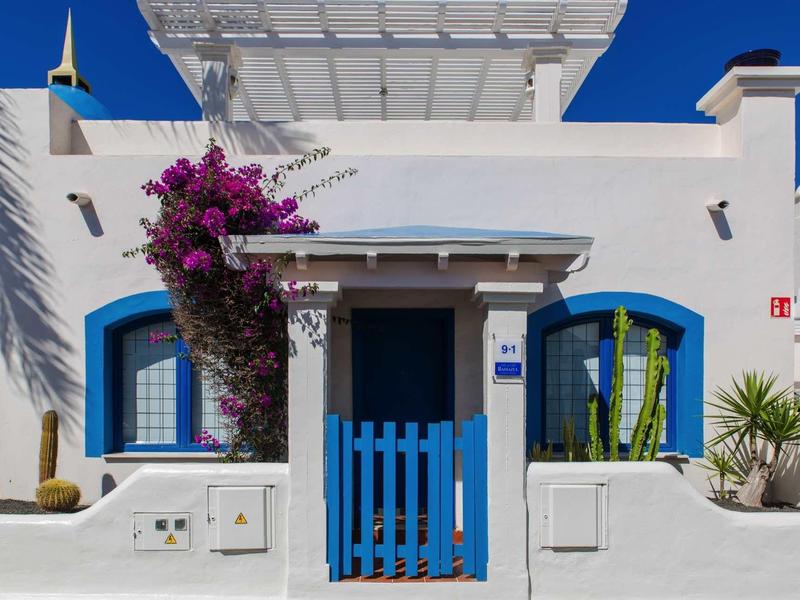 White house with blue door and windows, surrounded by flowers and clear sky.