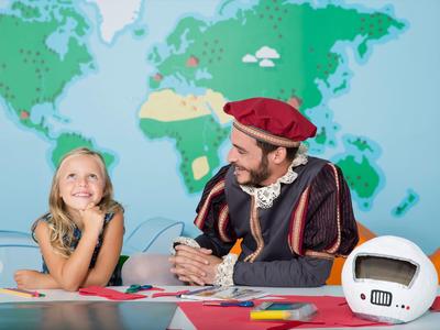 Child and man in costume sitting at table with world map background, space helmet on table.