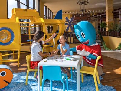Children and a mascot character sit at a table in a bright indoor play area with colorful decorations.