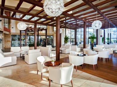 Spacious hotel lounge with white chairs, wooden beams, and large windows letting in natural light.