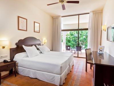 Bright hotel room with a comfortable bed, desk, TV, and balcony overlooking greenery.