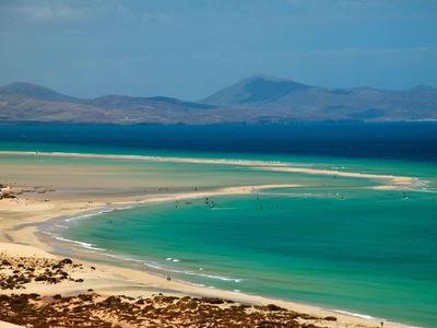 Curved sandy beach with turquoise water and distant mountains under a partly cloudy sky.