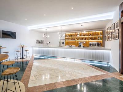 Modern hotel bar with illuminated counter, wooden stools, and marble flooring under bright lighting.