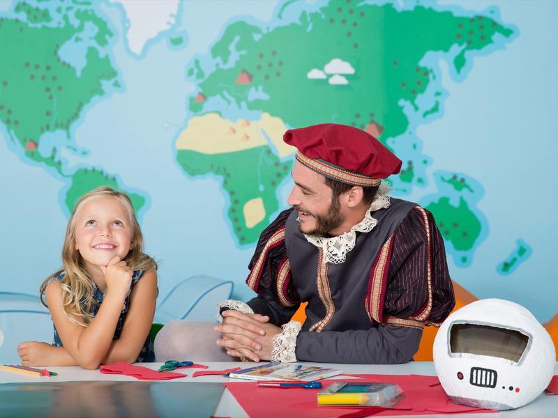 Child and man in costume sitting at table with world map background, space helmet on table.