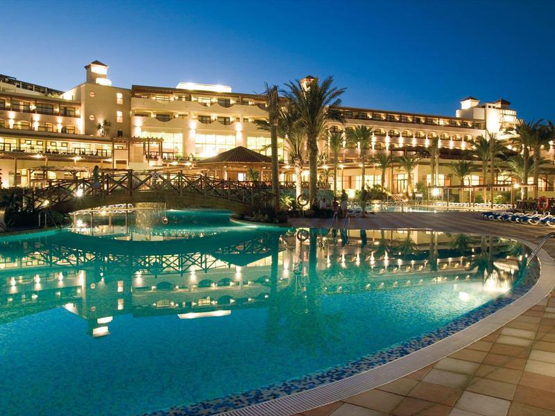 Luxurious hotel illuminated at night with large outdoor pool and palm trees.