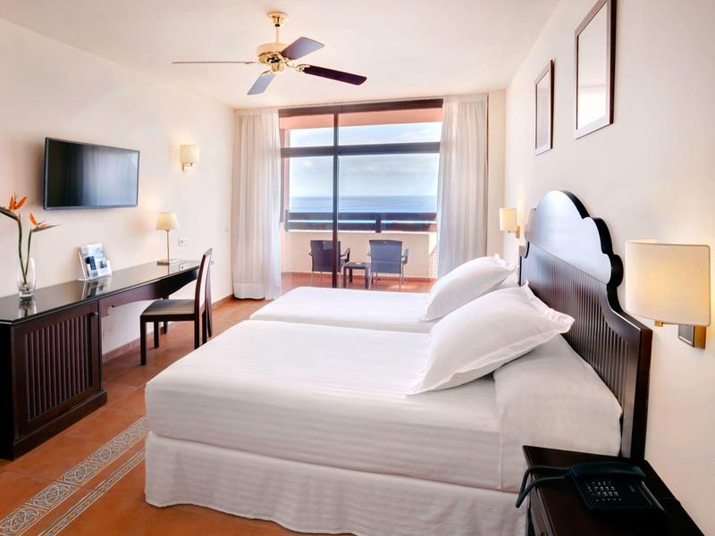 Bright hotel room with a large bed, ocean view, desk, and balcony seating.