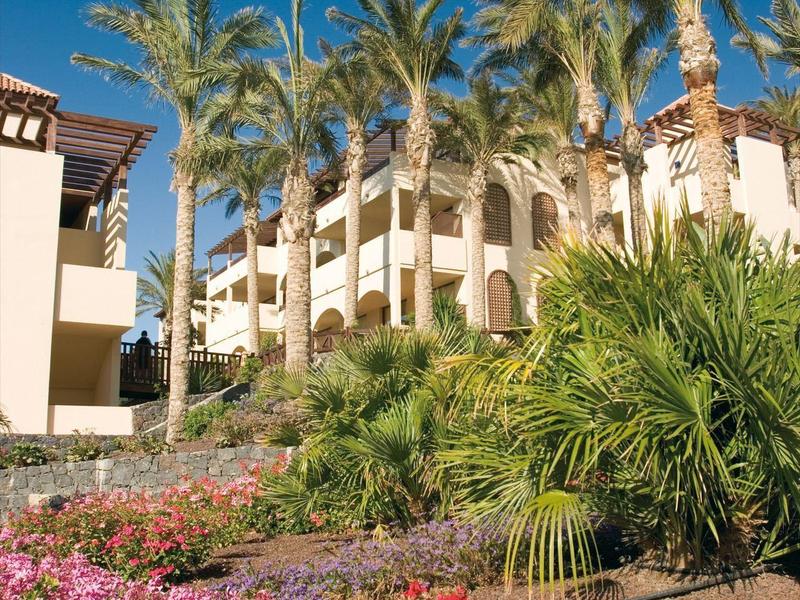 Resort with palm trees, pink flowers, and beige buildings under a clear sky.