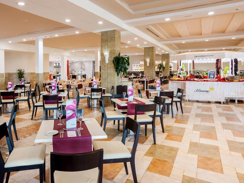 Spacious hotel dining area with tables, chairs, and buffet setup under bright lighting.