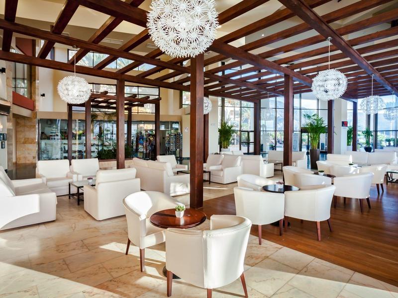Spacious hotel lounge with white chairs, wooden beams, and large windows letting in natural light.