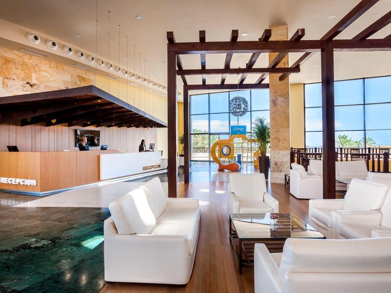 Modern hotel lobby with white sofas, wooden flooring, and large glass windows with ocean view.
