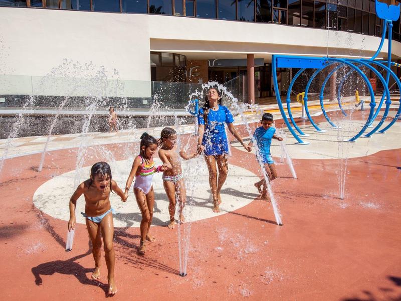 Children playing and running through outdoor water fountains on a sunny day.
