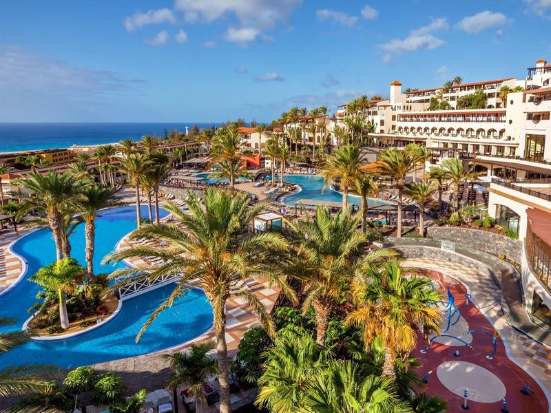Resort with multiple pools, palm trees, lounge areas, and ocean view under a blue sky.