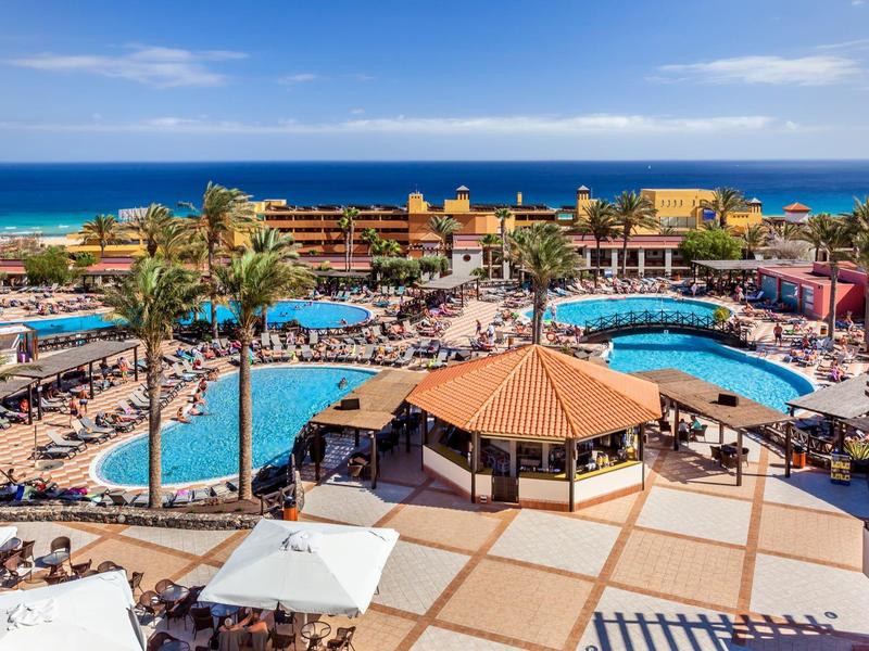 Large resort pool area with palm trees and buildings by the ocean under a blue sky.