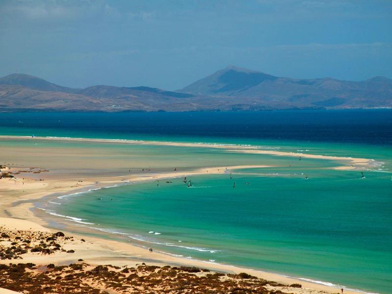 Curved sandy beach with turquoise water and distant mountains under a partly cloudy sky.