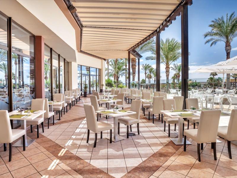 Spacious outdoor hotel restaurant terrace with tables, chairs, palm trees, and sunny weather.