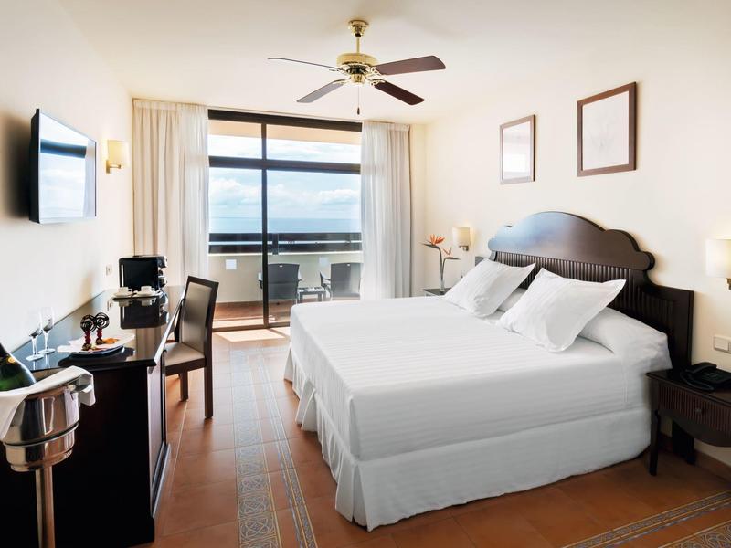 Bright hotel room with twin beds, ceiling fan, balcony with sea view, and modern amenities.