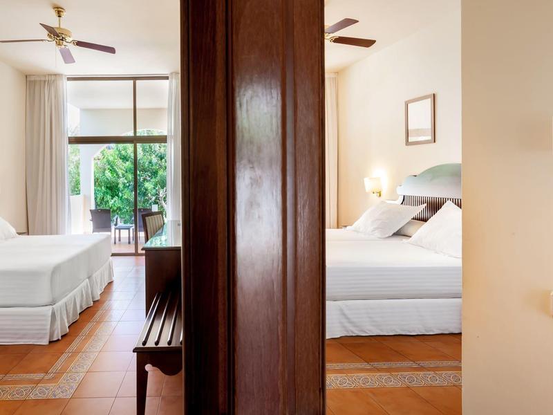 Two hotel rooms separated by a wooden wall, each with a bed, ceiling fan, and window with curtains.