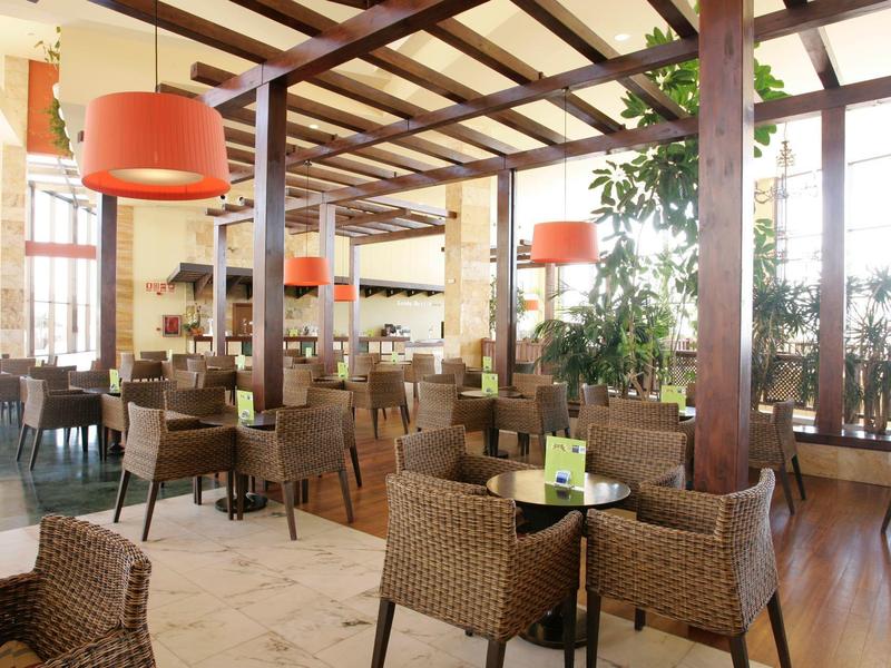 Spacious hotel dining area with wicker chairs, wooden beams, large windows, and orange pendant lights.
