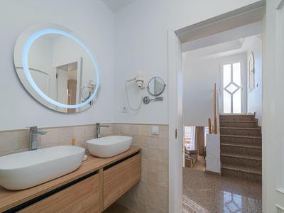 Modernes Badezimmer mit zwei weißen Waschbecken, Holzschrank, großem rundem Spiegel und Treppen im Hintergrund.