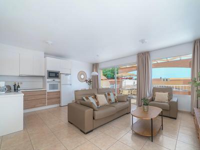 Helles Wohnzimmer mit brauem Sofa, zwei runden Tischen, offener Küche und großen Fenstern mit Blick aufs Meer.