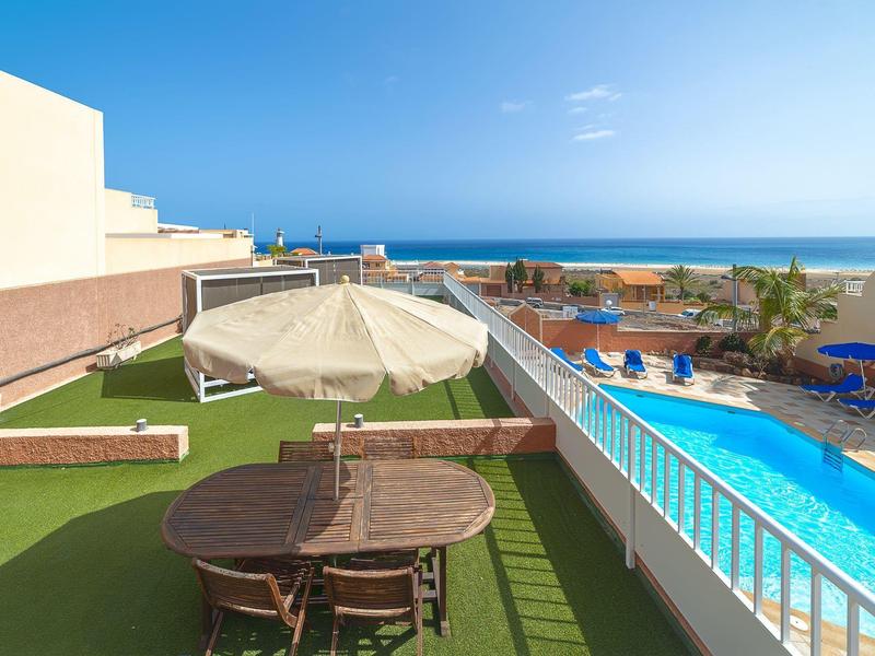 Vue d'une terrasse d'hôtel avec table, chaises, parasol et piscine avec vue sur la mer.
