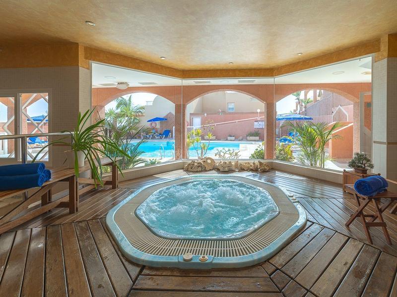 Espace intérieur confortable avec jacuzzi, sol en bois et vue sur la piscine extérieure et les chaises longues.