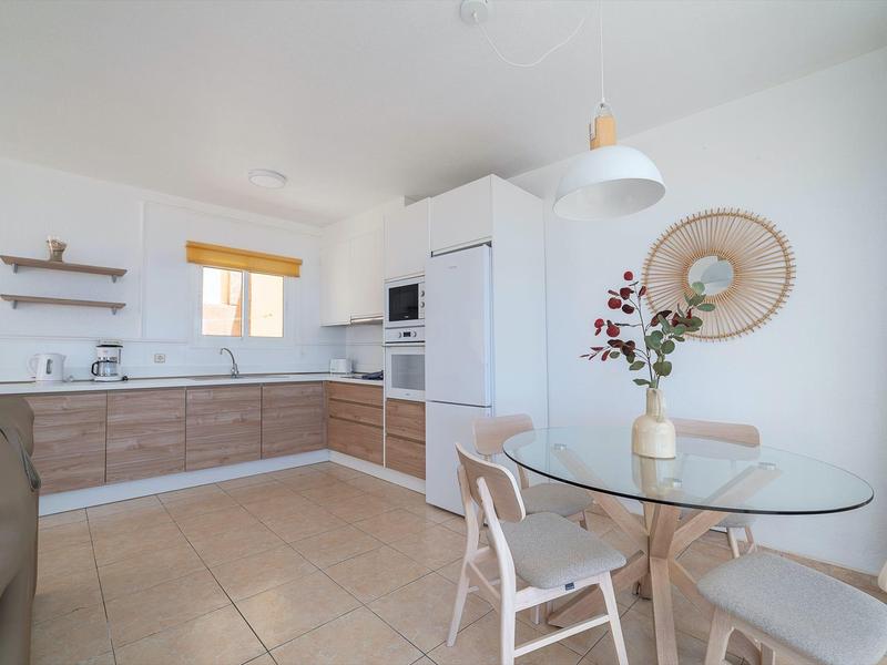 Cuisine moderne avec armoires en bois, table en verre et chaises dans un espace lumineux et minimaliste.