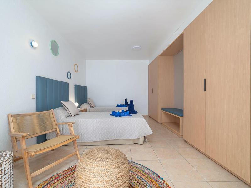 Chambre lumineuse avec lit double, chaise en bois, grande armoire et tapis sur sol carrelé.