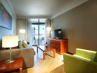 Chambre d'hôtel moderne avec canapé, fauteuil, télévision et balcon à la lumière du jour.