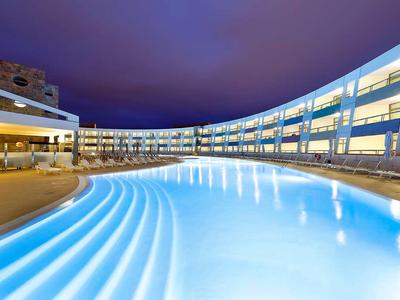 Nowoczesny hotel z dużym podświetlanym basenem na zewnątrz nocą.