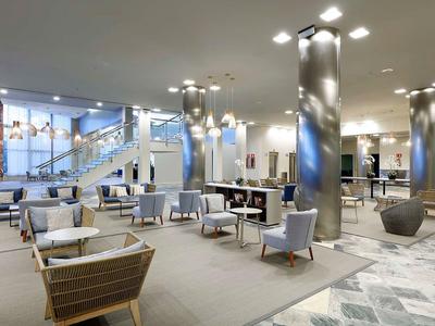 Przestronna nowoczesna przestrzeń wypoczynkowa hotelu z fotelami, stolikami i kolumnami w neutralnych kolorach.