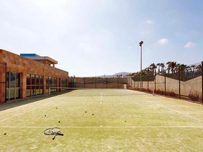 Pista de tenis vacía junto a un edificio con cercas y palmeras al fondo en un día soleado
